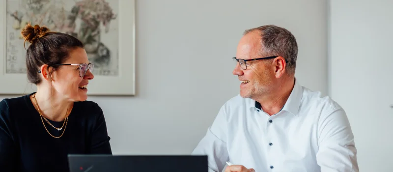 Zwei Personen sitzen an einem Tisch mit Laptop und sprechen miteinander. Das Bild steht für praxisnahen Austausch und Unternehmensberatung für Waren-, Vieh- und Dienstleistungsgenossenschaften.
