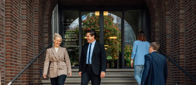 Mehrere Personen in Geschäftskleidung begegnen sich am Eingang eines Backsteingebäudes. Das Bild steht für praxisnahe Orientierung und hilfreiche Tipps für neue Genossenschaften.