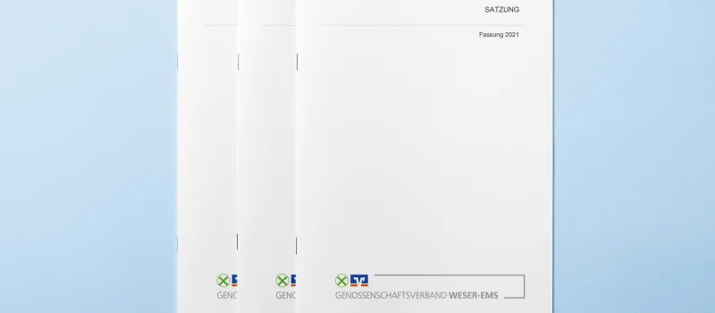 Mehrere Exemplare der Satzung des Genossenschaftsverbands Weser-Ems liegen auf hellblauem Untergrund. Das Bild steht für die Werte und rechtlichen Grundlagen des Verbands.