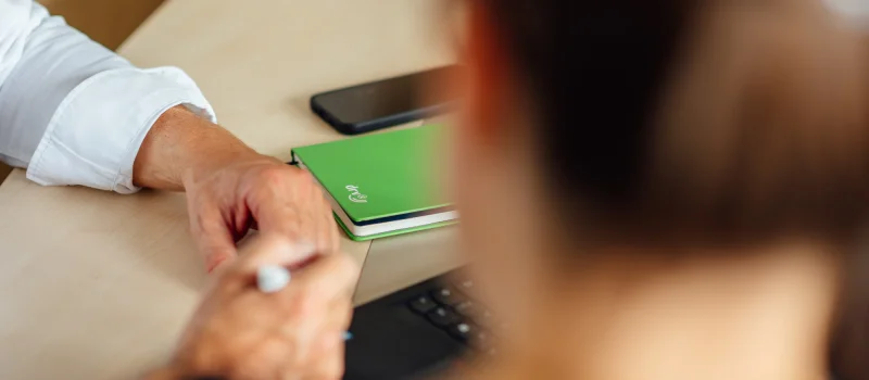 Eine Person hält einen Stift an einem Besprechungstisch; daneben liegen ein grünes Notizbuch und Smartphones. Das Bild steht für Abstimmung und Zusammenarbeit mit Bundes- und Regionalverbänden.