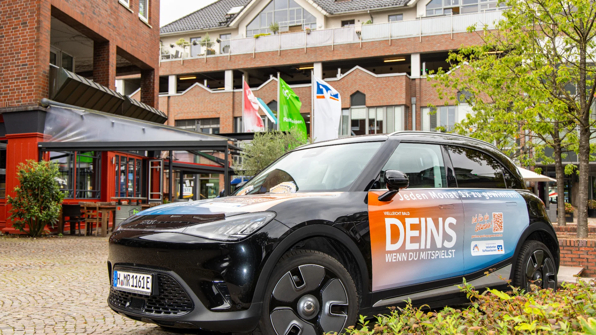 Ein beklebtes GewinnSpar-Auto mit dem Schriftzug „DEINS, wenn du mitspielst“ steht vor einem Gebäude mit Volksbank- und Raiffeisen-Flaggen. 