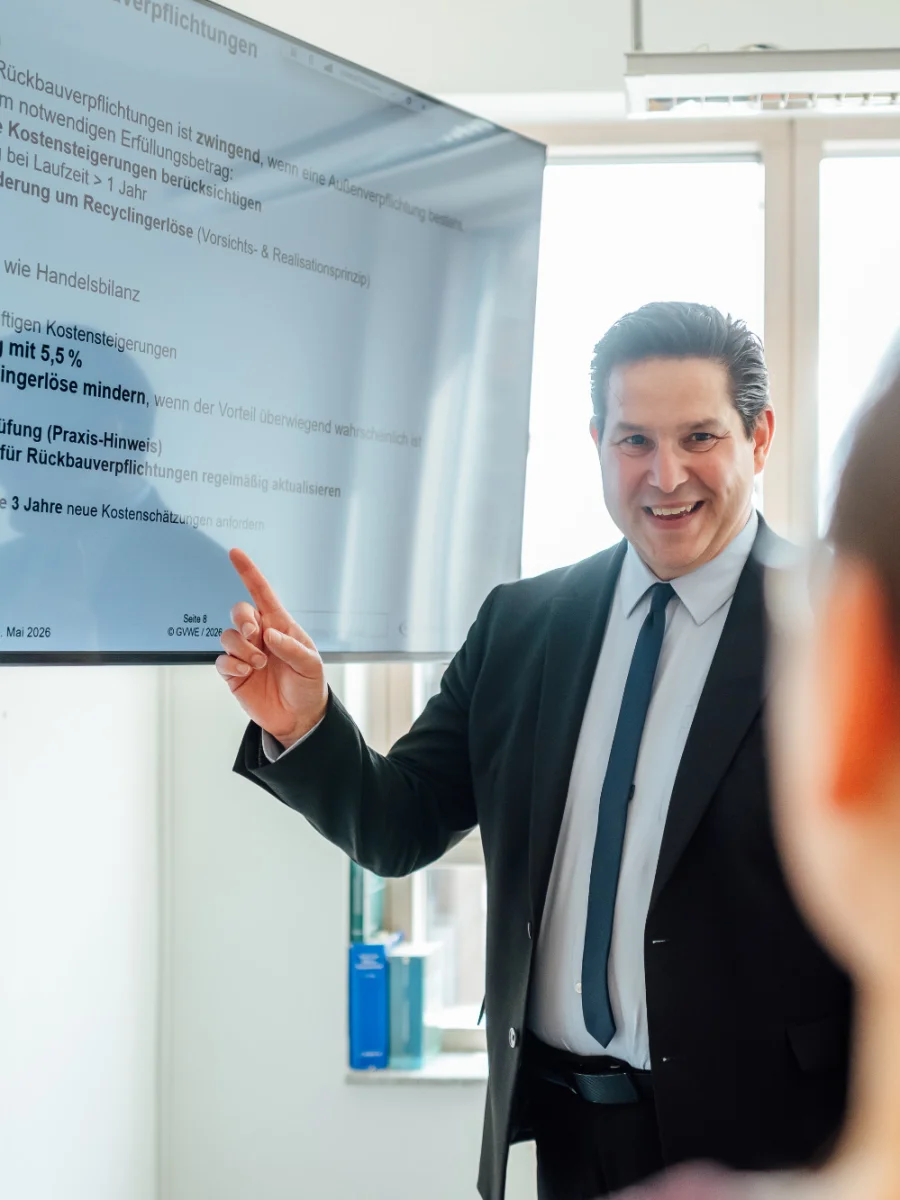 Ein Berater steht vor einem Bildschirm mit steuerlichen Stichpunkten und zeigt auf eine Präsentation. Das Bild steht für steuerliche Orientierung und fachliche Steuerberatung beim Genossenschaftsverband Weser-Ems.