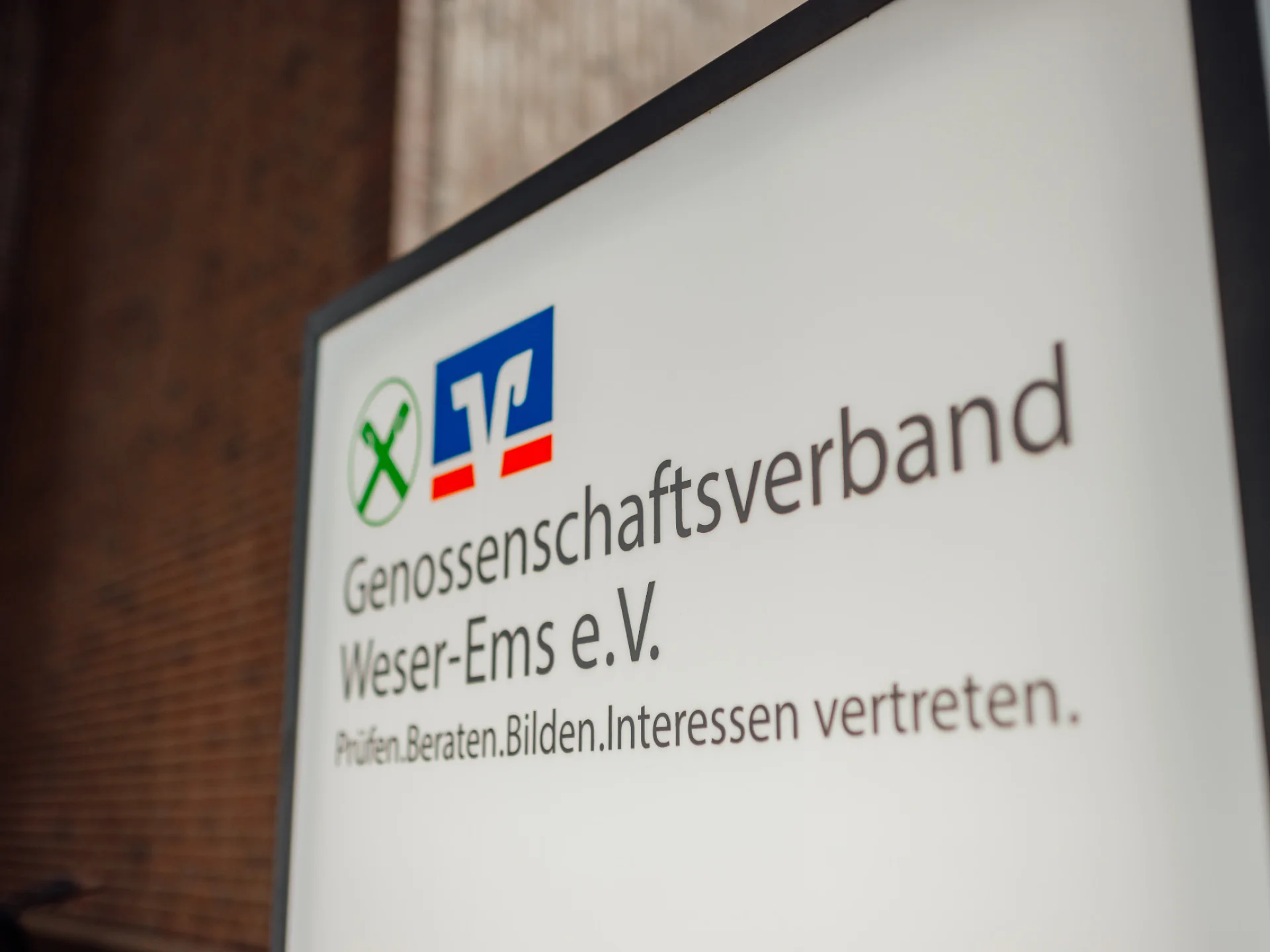 Schild des Genossenschaftsverbands Weser-Ems mit Logo und den Begriffen Prüfen, Beraten, Bilden und Interessen vertreten. Das Bild begleitet die Seite zu den Jahres-, Nachhaltigkeits- und Transparenzberichten des GVWE.