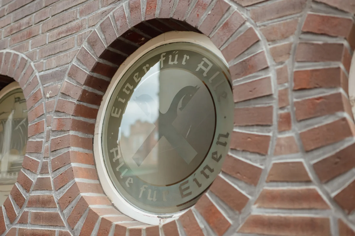 Rundes Fenster in einer Backsteinwand mit Raiffeisen-Symbol und dem Schriftzug „Einer für Alle, Alle für Einen“.
