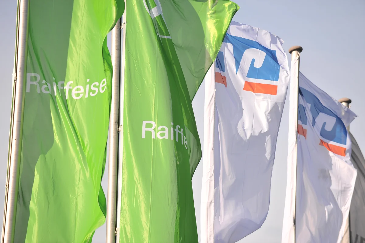 Grüne Raiffeisen-Flaggen und weiße Flaggen mit VR-Logo wehen vor hellem Himmel.