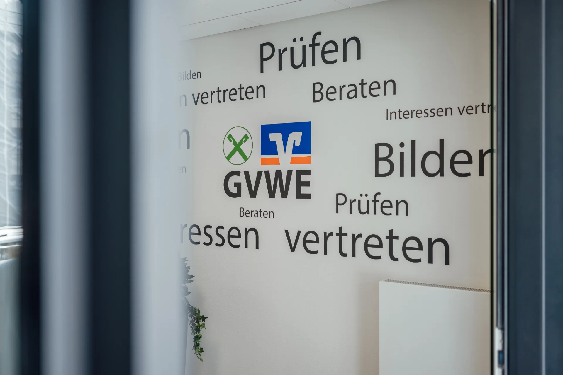 Blick durch eine Glastür auf eine Wand mit GVWE-Logo und Begriffen wie Prüfen, Beraten, Bilden und Interessen vertreten. Das Bild begleitet die Seite zu den Mitgliedsgenossenschaften des GVWE.
