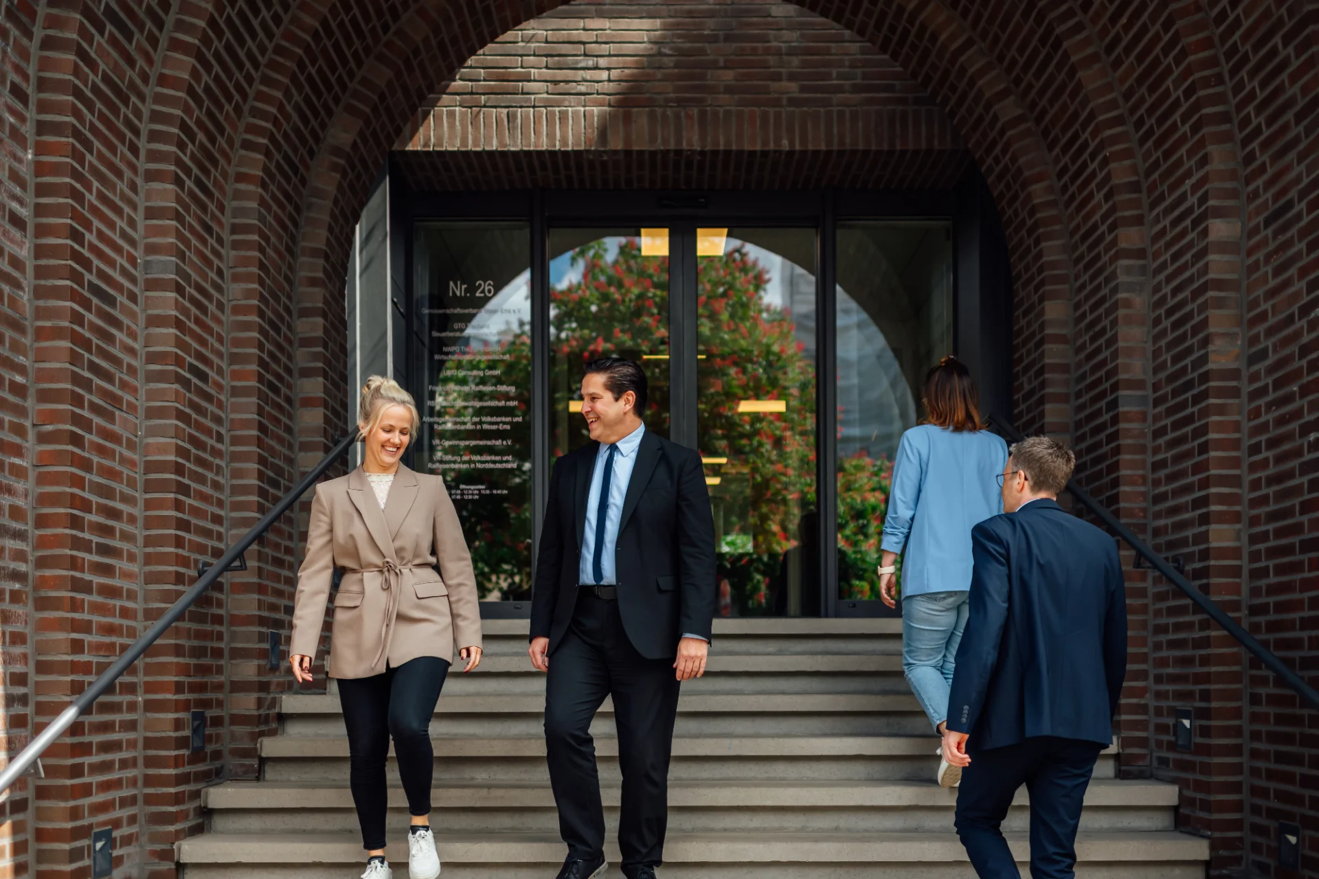 Mehrere Personen in Geschäftskleidung begegnen sich am Eingang eines Backsteingebäudes. Das Bild steht für praxisnahe Orientierung und hilfreiche Tipps für neue Genossenschaften.