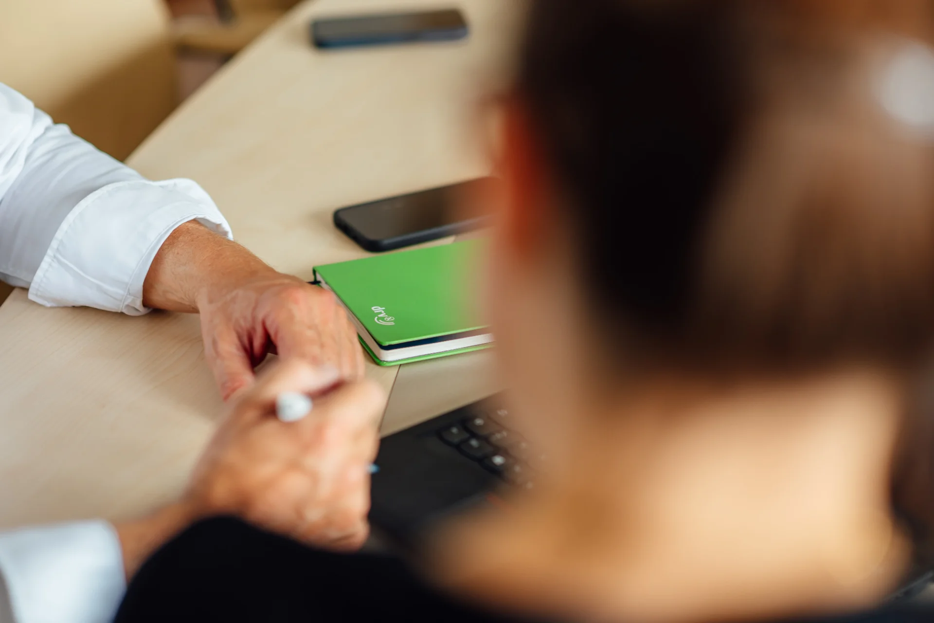 Eine Person hält einen Stift an einem Besprechungstisch; daneben liegen ein grünes Notizbuch und Smartphones. Das Bild steht für Abstimmung und Zusammenarbeit mit Bundes- und Regionalverbänden.